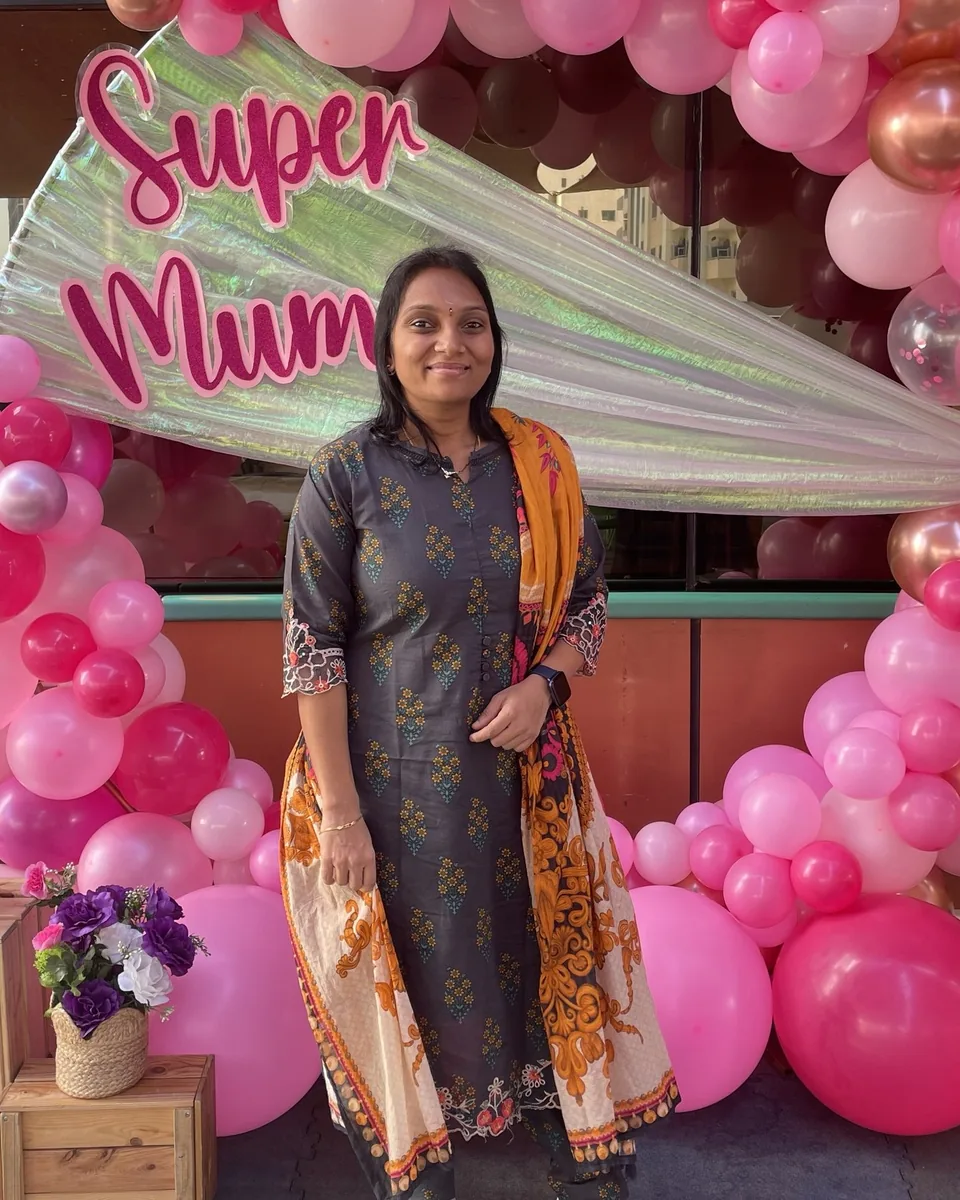 Sowmya Boga smiling in front of a pink balloon arch with a Super Mum sign